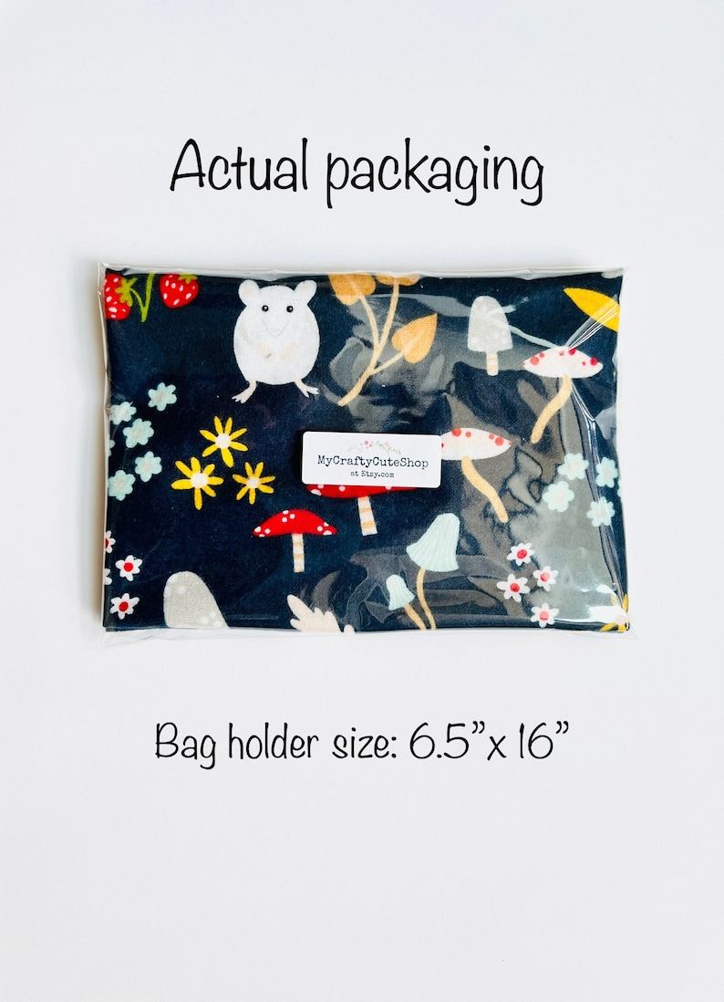 Op de afbeelding: Een doorzichtige plastic zak met een zwarte stoffen tas erin. De tas heeft een witte muis, rode en witte paddenstoelen en gele en groene bladeren op een donkerblauwe achtergrond. De zak is gelabeld met "MyCraftyCuteShop at Etsy.com". De maat van de tasdrager is 16,5 cm x 40,6 cm.