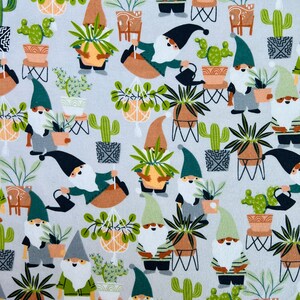 Op de afbeelding: Een witte stof met een herhalend patroon van gnomen die potplanten vasthouden. De gnomen dragen groene hoeden en hebben witte baarden. De planten zijn een verscheidenheid aan cactussen, vetplanten en hangplanten. De achtergrond is lichtgrijs.