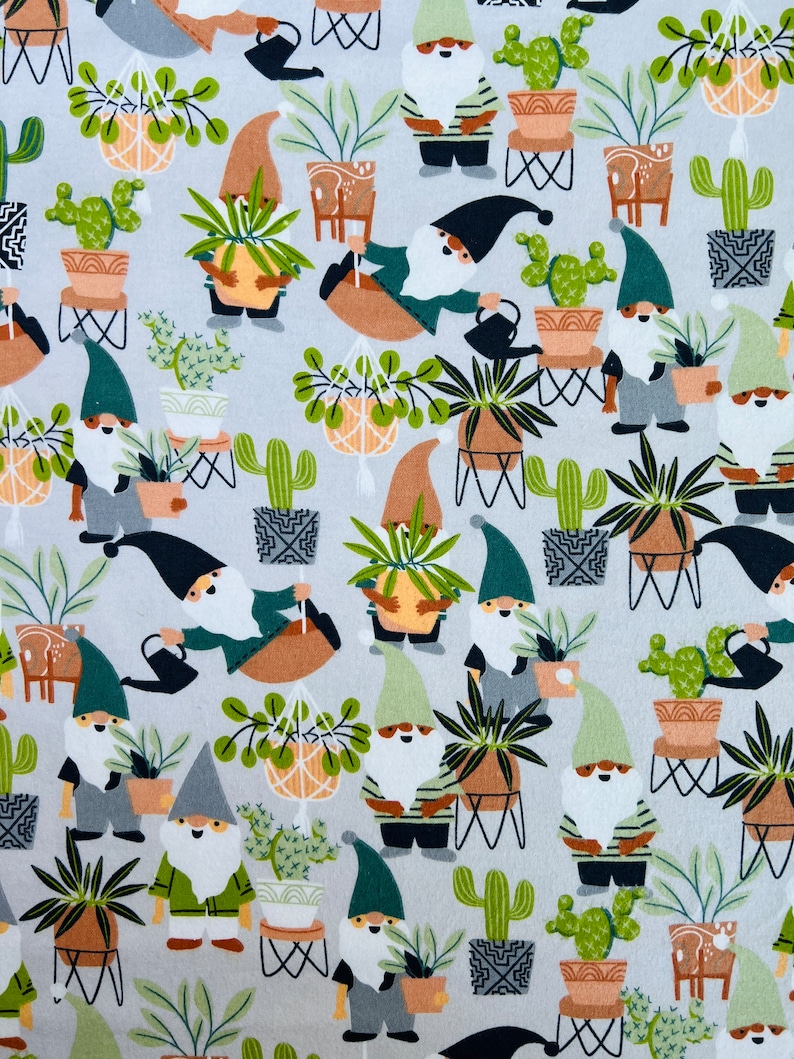 Op de afbeelding: Een witte stof met een herhalend patroon van gnomen die potplanten vasthouden. De gnomen dragen groene hoeden en hebben witte baarden. De planten zijn een verscheidenheid aan cactussen, vetplanten en hangplanten. De achtergrond is lichtgrijs.