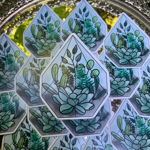 以下が含まれることがあります： 緑色の植物と多肉植物が描かれた幾何学的なテラリウムのデザインのステッカー10枚セット。ステッカーは、青い色合いの白い背景に印刷されています。