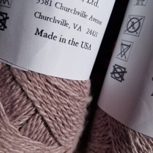Op de afbeelding: Twee bollen garen in een stoffige roze kleur. Het etiket vermeldt "Cestari, Ltd. 3581 Churchville Avenue Churchville, VA 24421 Made in the USA". Het etiket bevat ook wasinstructies.