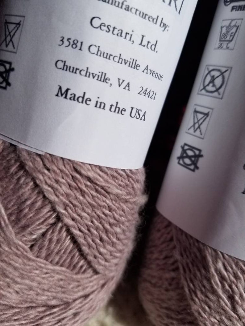 Op de afbeelding: Twee bollen garen in een stoffige roze kleur. Het etiket vermeldt "Cestari, Ltd. 3581 Churchville Avenue Churchville, VA 24421 Made in the USA". Het etiket bevat ook wasinstructies.
