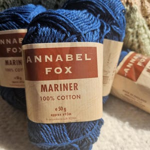 May include: Close-up of several skeins of blue Annabel Fox Mariner 100% cotton yarn. Each skein is labeled with the brand name, yarn type, and weight of 50g. The yarn is a deep, rich blue color.