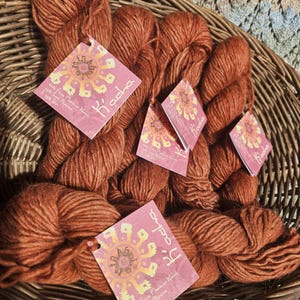 May include: A basket filled with rust-colored yarn skeins. Each skein has a pink tag with the word "K'acha" and a decorative design. The yarn is made of 60% Fine Merino Wool and 40% Silk.