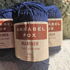 May include: Three spools of navy blue Annabel Fox Mariner 100% cotton yarn. Each spool has a label with the brand name and product details. The yarn is tightly wound, and the labels are a mix of red and cream colors.