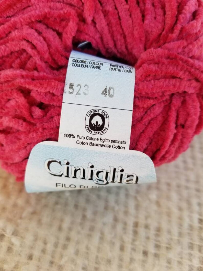 Pu&ograve; includere: Un gomitolo di filato rosa acceso con un'etichetta bianca che recita "COLORE/COLOUR COULUER/FARBE PARTITA/LOT PARTIE/BAIN 1523 40 COTONE 100% FIBRA NATURALE 100% Puro Cotone Egitto pettinato Coton Baumwolle Cotton Ciniglia FILO DI"