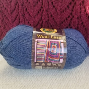 Peut inclure: Une pelote de laine bleue avec une étiquette « Wool-Ease » est présentée. L'étiquette présente un motif de crochet coloré. L'arrière-plan montre un tissu tricoté bordeaux.