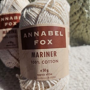 May include: Close-up of two cream-colored yarn skeins. The labels read "ANNABEL FOX MARINER 100% COTTON e 50g approx e93m". The yarn is tightly wound, and the labels are a dark brown with white text. The background is a soft, out-of-focus green.
