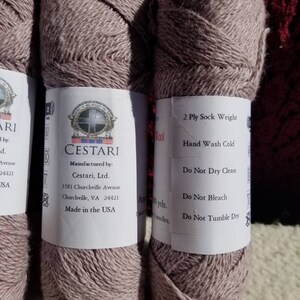 Op de afbeelding: Twee bollen lichtbruin garen, met het label "CESTARI" en "2 Ply Sock Weight". Het garen is gemaakt in de VS en is machinewasbaar.