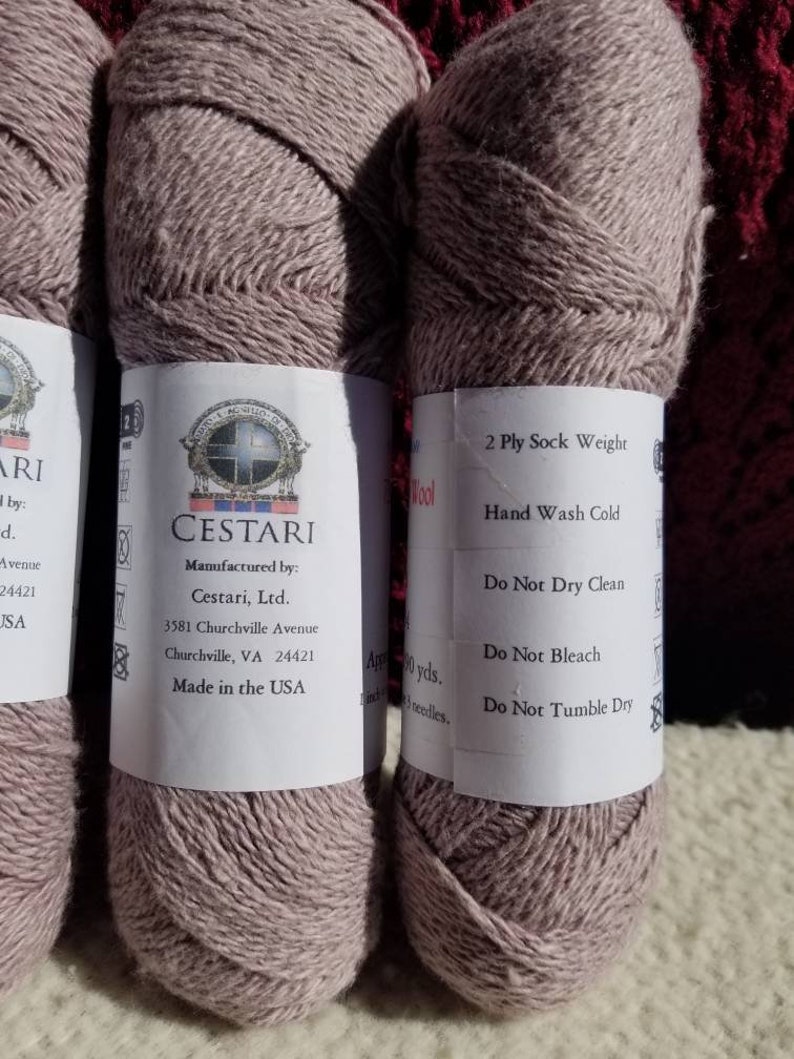 Op de afbeelding: Twee bollen lichtbruin garen, met het label "CESTARI" en "2 Ply Sock Weight". Het garen is gemaakt in de VS en is machinewasbaar.