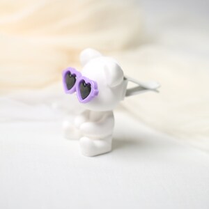 May include: A white figurine of a bear wearing heart-shaped sunglasses with purple frames and black lenses. The bear is sitting down. The background is a soft, blurred white and cream color.