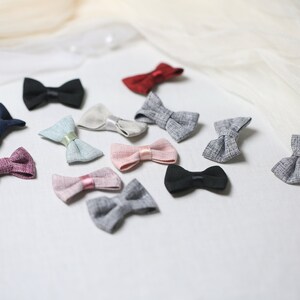 May include: A collection of small, fabric bow ties in various colors, including black, navy, red, pink, and gray. Some bows have satin ribbon accents. The bows are arranged on a white surface with a soft, draped fabric in the background.