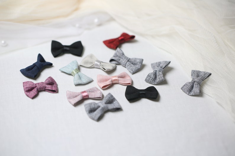 May include: A collection of small, fabric bow ties in various colors, including black, navy, red, pink, and gray. Some bows have satin ribbon accents. The bows are arranged on a white surface with a soft, draped fabric in the background.