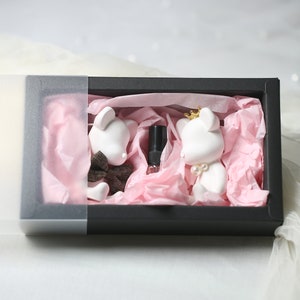 May include: A black gift box with a clear lid, containing two white bear figurines and a small bottle. The bears are decorated with a bow tie, a crown, and pearls. The box is lined with pink tissue paper, creating a charming presentation.
