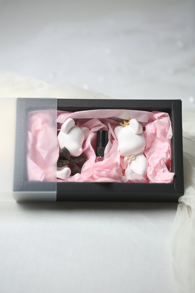 May include: A black gift box with a clear lid, containing two white bear figurines and a small bottle. The bears are decorated with a bow tie, a crown, and pearls. The box is lined with pink tissue paper, creating a charming presentation.