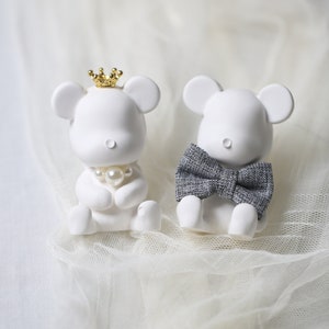 May include: Two white mouse figurines. One wears a gold crown and a pearl necklace, the other a gray bow tie. The figurines are sitting on a white, textured surface.