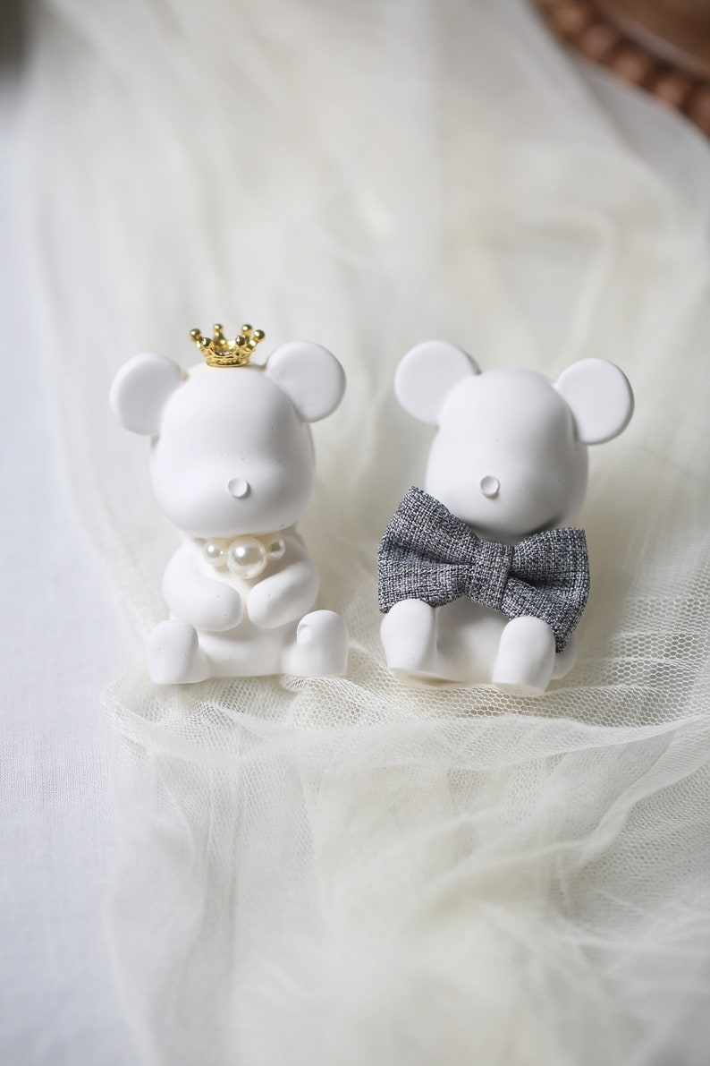 May include: Two white mouse figurines. One wears a gold crown and a pearl necklace, the other a gray bow tie. The figurines are sitting on a white, textured surface.