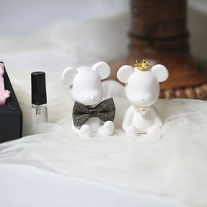 May include: Two white bear figurines, one with a bow tie and the other with a crown and pearl necklace. A small perfume bottle and a black box with pink tissue paper are also in the image. The figurines are on a white, textured surface.