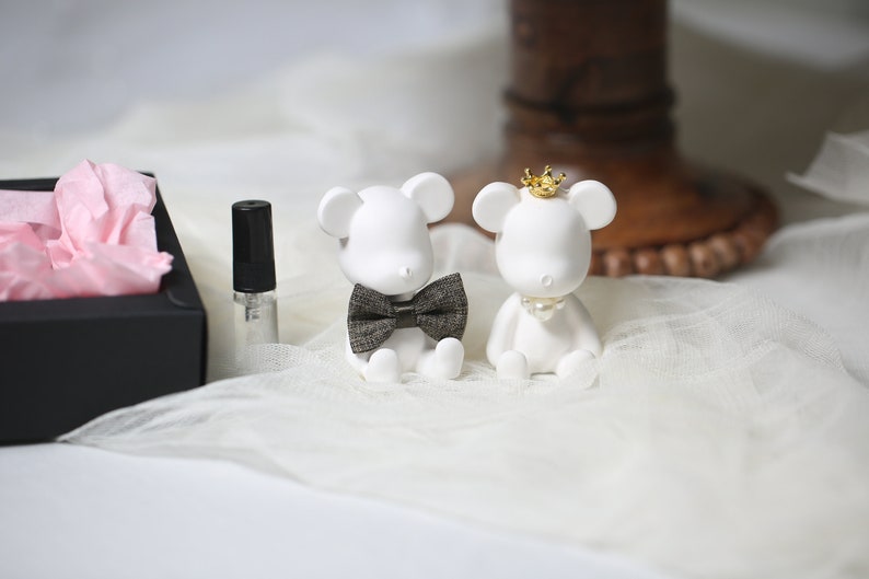 May include: Two white bear figurines, one with a bow tie and the other with a crown and pearl necklace. A small perfume bottle and a black box with pink tissue paper are also in the image. The figurines are on a white, textured surface.
