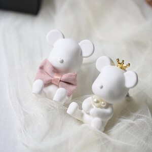 May include: Two white mouse figurines. One mouse wears a pink bow tie, the other a gold crown and pearl necklace. The figurines are sitting on a white, sheer fabric.