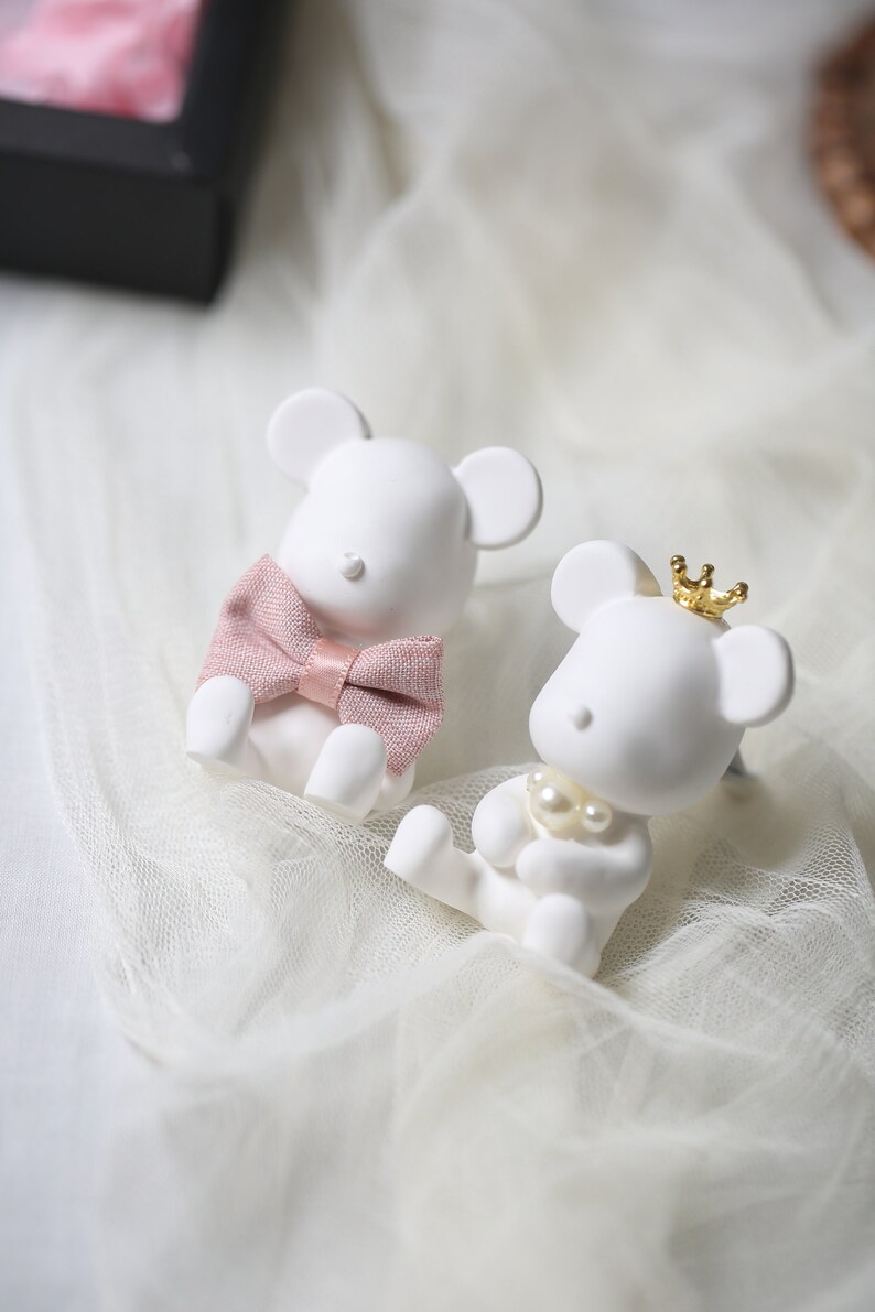 May include: Two white mouse figurines. One mouse wears a pink bow tie, the other a gold crown and pearl necklace. The figurines are sitting on a white, sheer fabric.