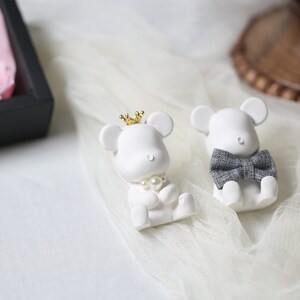 May include: Two white mouse figurines, one with a gold crown and pearl necklace, the other with a gray bow tie. They are sitting on a white, sheer fabric. A black box with pink tissue paper is in the background.