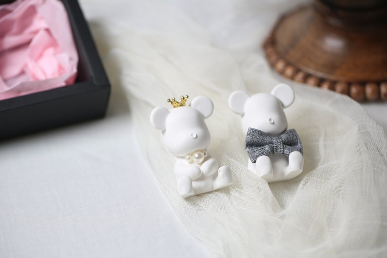 May include: Two white mouse figurines, one with a gold crown and pearl necklace, the other with a gray bow tie. They are sitting on a white, sheer fabric. A black box with pink tissue paper is in the background.