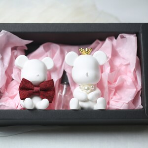 May include: Two white bear figurines, one with a red bow tie and the other with a gold crown and pearl necklace, sit in a black gift box with pink tissue paper. A small clear bottle is also in the box.