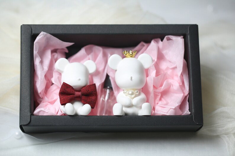 May include: Two white bear figurines, one with a red bow tie and the other with a gold crown and pearl necklace, sit in a black gift box with pink tissue paper. A small clear bottle is also in the box.