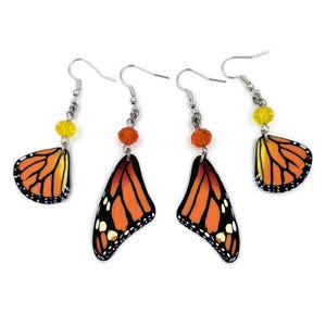 May include: Four pairs of butterfly wing earrings. The earrings feature orange and black wings with white spots, silver hooks, and yellow or orange beads. The wings are detailed with black outlines and veins.