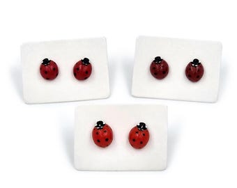 May include: Three pairs of ladybug stud earrings. Each earring is a red ladybug with black spots and a tiny black hat. The earrings are displayed on white rectangular cards against a white background.
