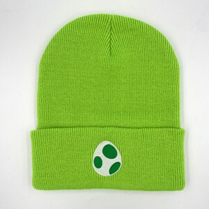 May include: A vibrant green knitted beanie hat featuring an embroidered egg design in white and green. The hat is made from a ribbed knit fabric. The egg design is on the front of the folded brim.