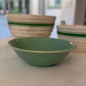 Peut inclure: Un bol en céramique vert clair au premier plan, avec un bord légèrement évasé. Derrière, deux grands bols crème avec des rayures vertes horizontales. Les bols sont probablement destinés à la cuisine.