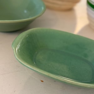 Może przedstawiać: Kolekcja zielonych, zabytkowych naczyń ceramicznych. Na pierwszym planie mały, owalny talerz z lekko podniesionym brzegiem. W tle widoczne są inne miski, prezentujące spójną paletę kolorów i design.