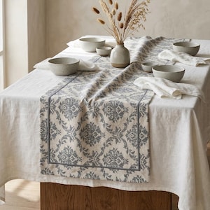 May include: A dining table set with a white tablecloth, a patterned table runner, and several bowls. The table runner features a gray and cream damask design. A vase with dried flowers sits in the center. Napkins and bowls are also on the table.