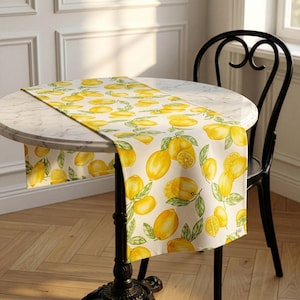May include: A yellow and white table runner with a lemon pattern. The runner is draped over a round marble-topped table. The table has a black metal base. A black chair is next to the table.