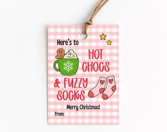 Pode incluir: Uma etiqueta de presente de Natal rosa e branca com o texto "Here's to Hot Chogs & Fuzzy Socks". A etiqueta apresenta ilustrações de uma caneca, um homem de gengibre e meias. É amarrada com barbante.