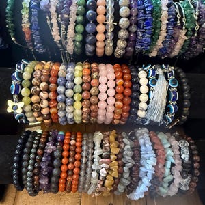 Könnte beinhalten: Eine Sammlung von Armbändern mit Perlen in verschiedenen Farben und Stilen. Die Armbänder verfügen über eine Vielzahl von Edelsteinen und Perlen, darunter Amethyst, Lapislazuli und Rosenquarz. Einige Armbänder haben Charms und Quasten.