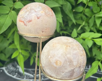 May include: Two white agate spheres with orange and brown inclusions are displayed on a gold metal stand. The spheres are smooth and polished.