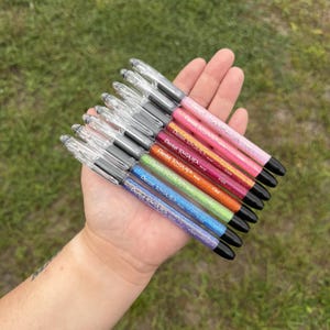 May include: A set of seven Pentel RSVP ballpoint pens with clear plastic caps and barrels filled with colorful glitter. The pens feature a fine point tip and are arranged in a fan shape, displaying shades of pink, orange, green, blue, and purple.
