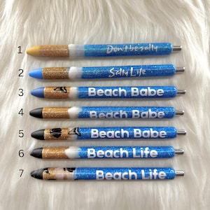 May include: A collection of seven pens with glittery blue barrels. The pens have various designs, including gold glitter accents and the phrases "Don't be salty," "Salty Life," "Beach Babe," and "Beach Life."