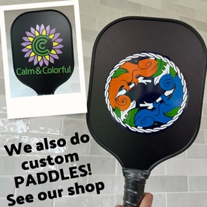 May include: Two black pickleball paddles. One paddle has a green, orange, and blue design with three stylized chameleons. The other paddle has a purple and green flower with the text "Calm & Colorful".