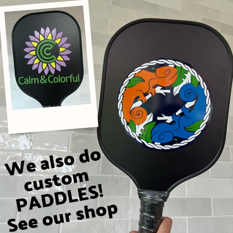 May include: Two black pickleball paddles. One paddle has a green, orange, and blue design with three stylized chameleons. The other paddle has a purple and green flower with the text "Calm & Colorful".