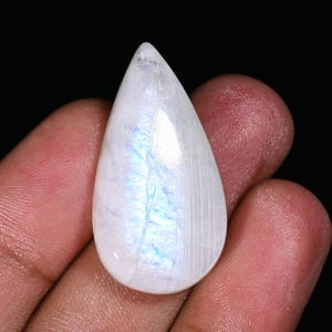 Puede incluir: Una piedra lunar pulida en forma de pera. La piedra es principalmente blanca con sutiles destellos azules e iridiscentes. La superficie es lisa y reflectante. La piedra preciosa se sostiene sobre un fondo oscuro.