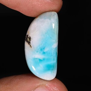 Può includere: Una gemma lucida di forma ovale con un'accattivante miscela di colori. La pietra presenta un gradiente di bianco, azzurro e turchese, con una piccola inclusione scura. La gemma è tenuta tra due dita su sfondo nero.