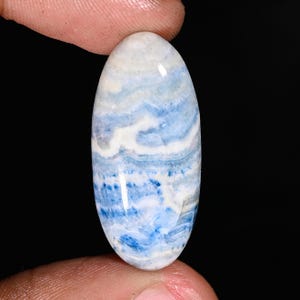 May include: An oval-shaped gemstone with a polished surface. The stone features a layered pattern of white, light blue, and deep blue hues. The gemstone is held between two fingers against a black background.