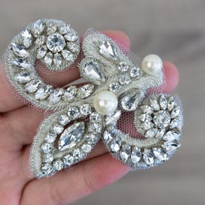 May include: A silver and white beaded applique with a pearl embellishment. The applique is made of a delicate fabric and is perfect for adding a touch of sparkle to any project.