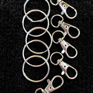 May include: A collection of silver-colored key rings and lobster clasp keychains. The key rings are round and of varying sizes. The lobster clasps have a swivel design and a small ring for attaching keys or other items. The items are arranged on a black surface.