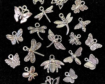 Könnte beinhalten: Eine Sammlung silberfarbener Anhänger in verschiedenen Insektenmotiven, darunter Schmetterlinge, Libellen und Bienen. Jeder Anhänger hat eine kleine Schlaufe zum Befestigen an Schmuck oder anderen Bastelarbeiten. Auf schwarzem Hintergrund.