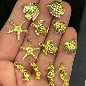May include: A collection of gold-colored charms featuring various sea creatures. The charms include starfish, turtles, fish, seahorses, mermaids, and scallop shells. Each charm has a small loop for attaching to jewelry or other crafts.